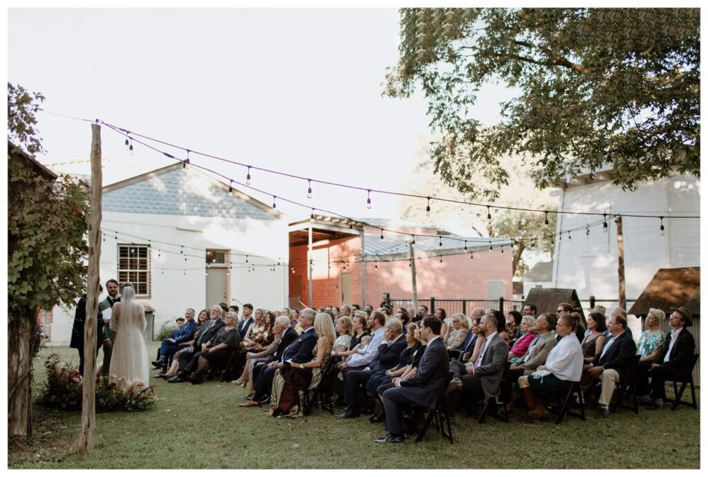An outdoor ceremony at an Ingenhuett on High wedding in Comfort, Texas, with guests seated under twinkling string lights as the couple exchanges vows in front of a cozy, historic backdrop.