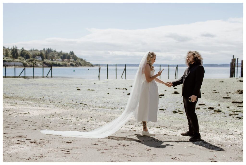 Bride and groom sharing a Ballard first look on a quiet beach before their Discovery Park wedding in Seattle, WA. The bride, in a tea-length dress and sneakers, reads her vows while holding hands with the groom, set against the serene waterfront. 