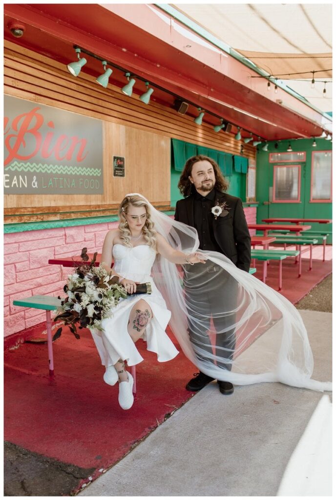 Bride and groom posing outside Un Bien, a vibrant Caribbean restaurant, before their Discovery Park wedding in Seattle, WA. The bride sits on a colorful picnic bench with her bouquet, her veil flowing in the breeze, while the groom stands beside her in a sleek black suit. A fun and stylish stop on their wedding day, filled with personality and bold colors.