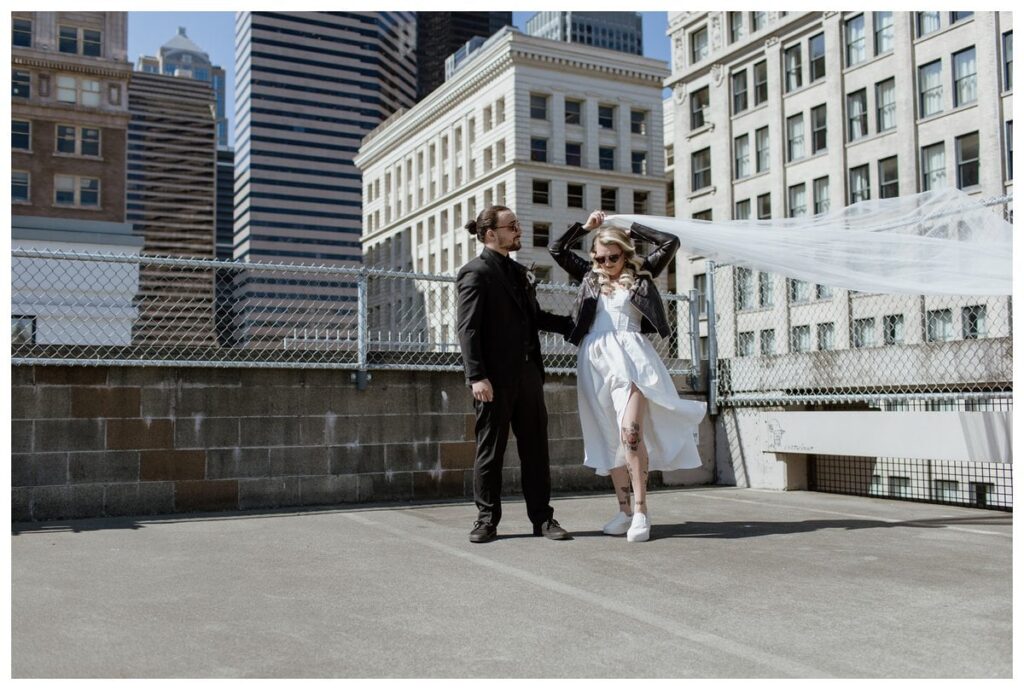 Bride and groom share a stylish moment on a Pioneer Square parking garage rooftop before their Discovery Park wedding in Seattle. The bride, rocking a leather jacket and sunglasses, lifts her veil in the wind, while the groom, dressed in all black, looks on against a backdrop of historic and modern city buildings. A bold, urban twist on their wedding day portraits.