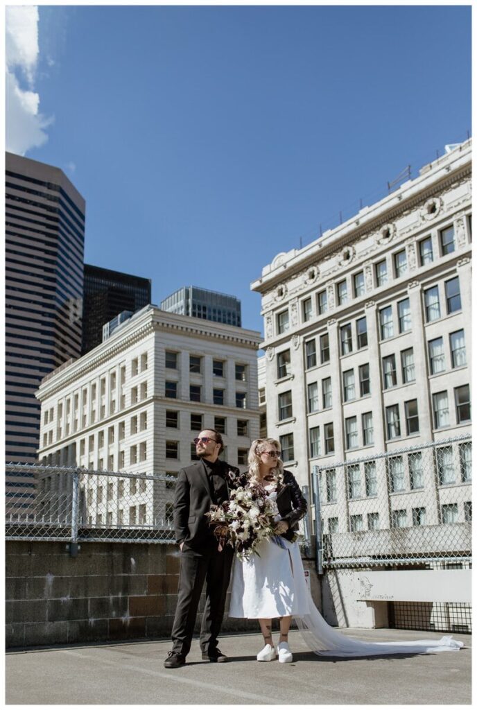 Bride and groom strike a stylish pose on a Pioneer Square parking garage rooftop before their Discovery Park wedding in Seattle. The couple, dressed in edgy wedding attire with sunglasses and a leather jacket, stand confidently against a backdrop of historic architecture and modern skyscrapers, creating a bold and unique urban wedding portrait.