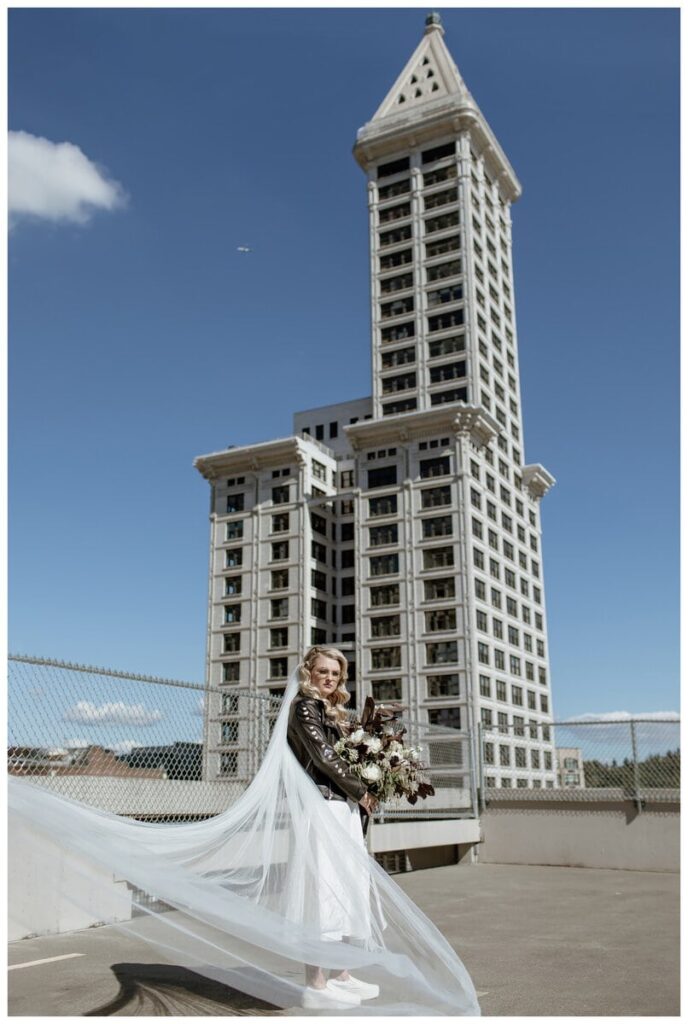 A bride stands on a Pioneer Square parking garage rooftop in Seattle, with her long veil flowing dramatically in the wind. Dressed in a white dress and black leather jacket, she holds a stunning bouquet against the backdrop of the iconic Smith Tower, creating a bold and stylish urban wedding portrait.