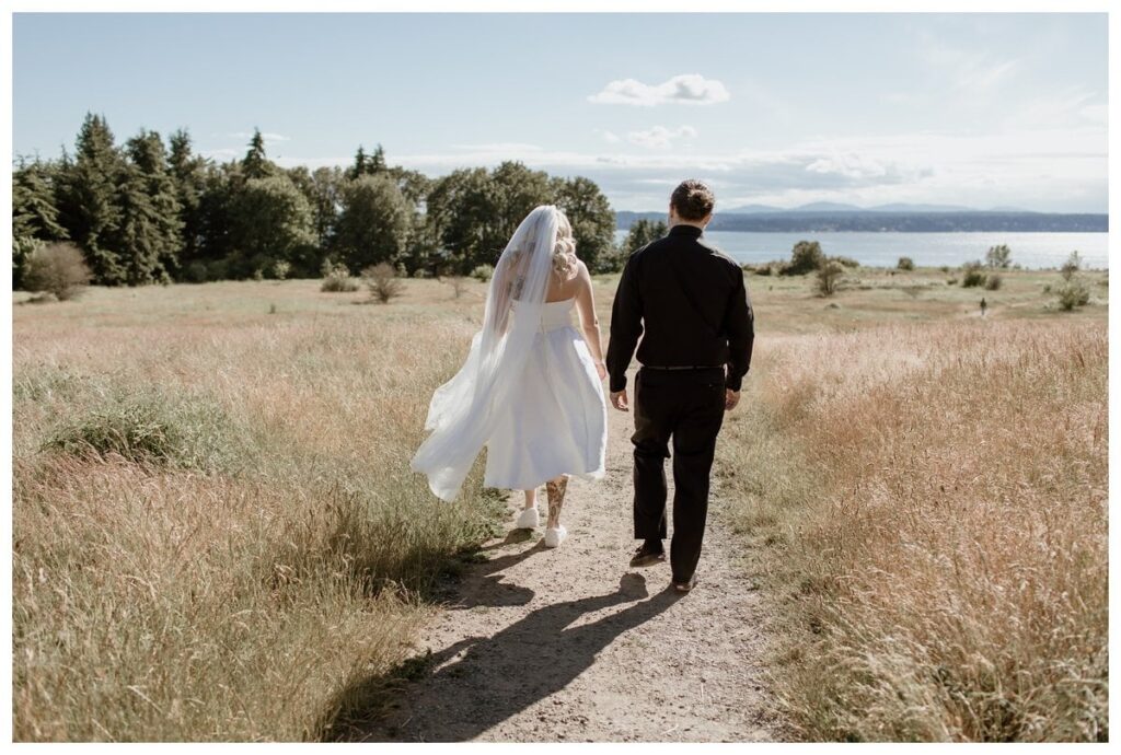 A bride and groom walk down a sunlit trail at their Discovery Park wedding in Seattle, surrounded by golden fields and a stunning waterfront view.