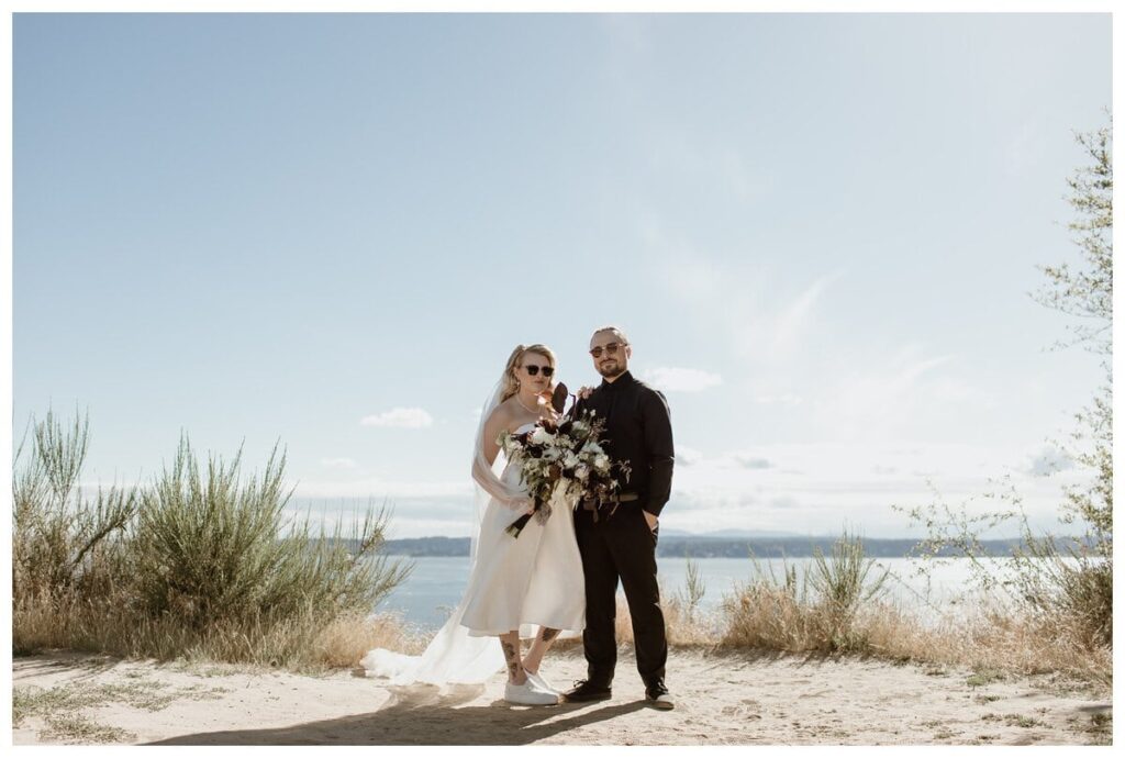 Bride and groom posing at their Discovery Park wedding in Seattle, WA, with a backdrop of Puget Sound and the Olympic Mountains. The bride wears a chic tea-length dress with sneakers and sunglasses, holding a lush bouquet, while the groom sports an all-black outfit. A stylish, intimate celebration in a natural setting.