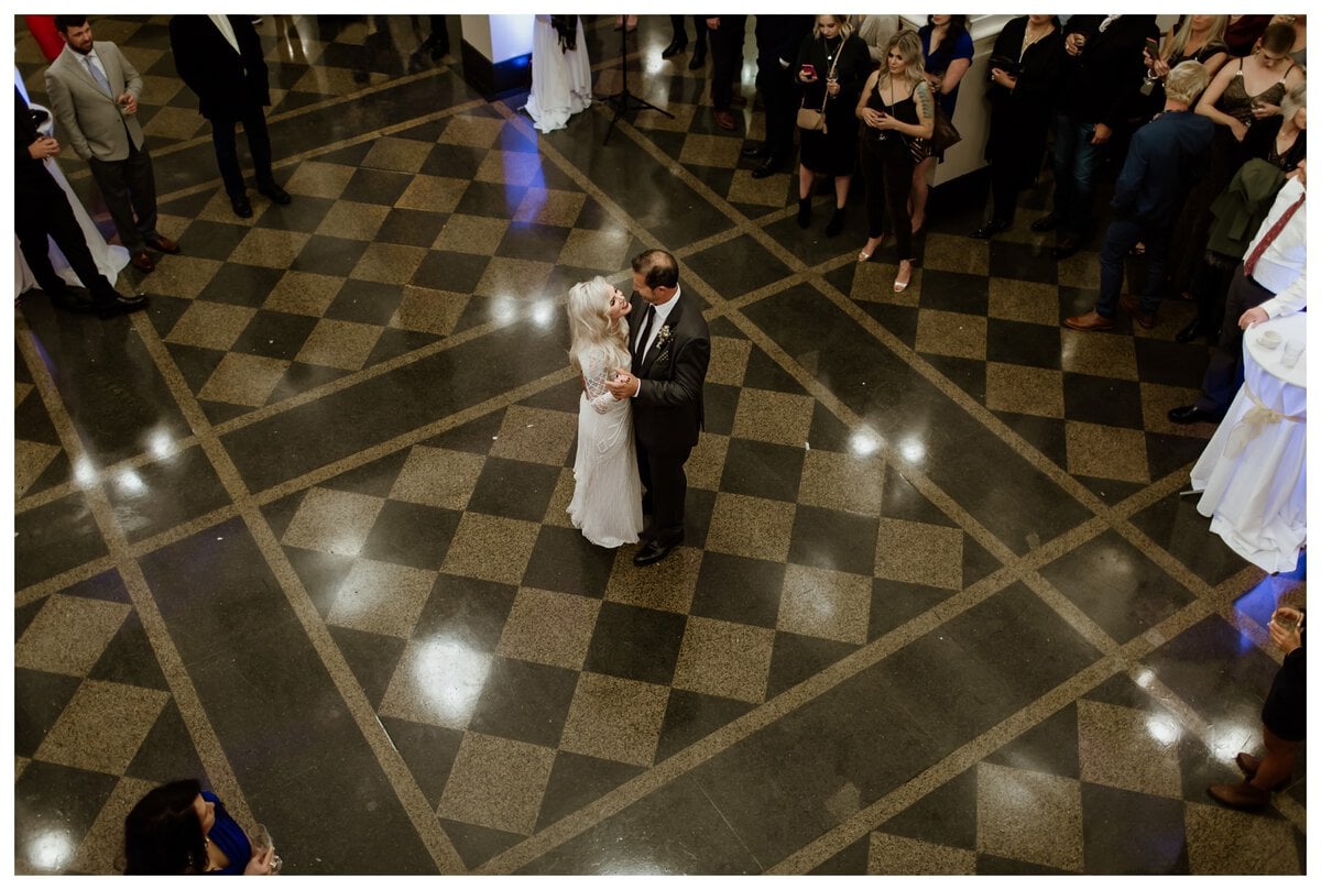 An overhead shot of a bride and groom sharing their first dance on a checkered ballroom floor, surrounded by wedding guests. A perfect moment for couples looking for cool first dance songs to make their wedding unique.