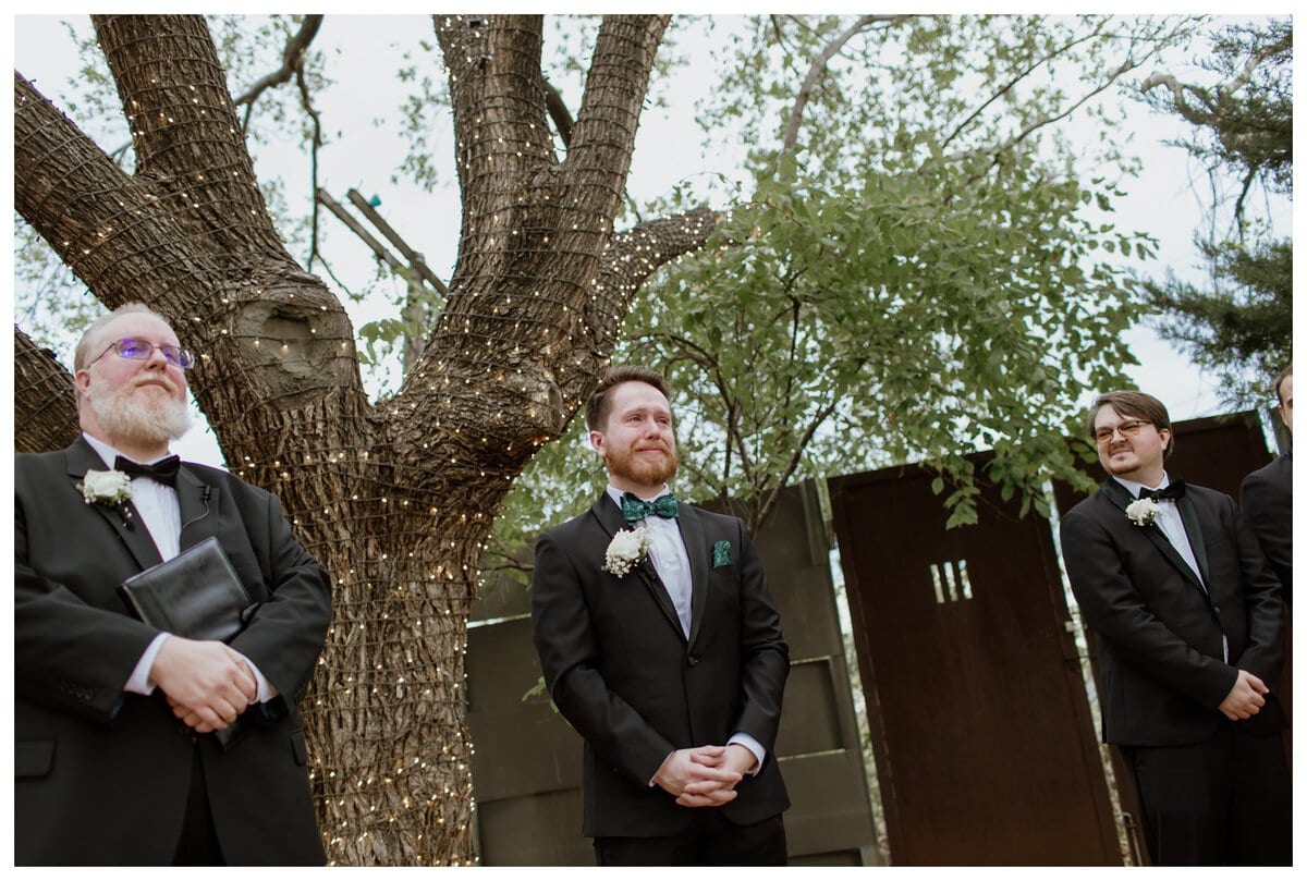 The groom stands at the altar with an emotional smile as he awaits his bride at an Artspace111 Fort Worth wedding. Twinkling string lights wrap around a large tree, creating a romantic outdoor setting. 