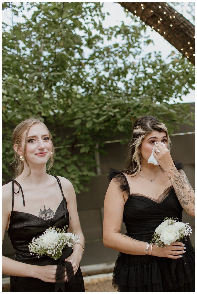 Emotional bridesmaids at an Artspace111 Fort Worth wedding, holding white rose bouquets. One bridesmaid wipes away tears while the other smiles with emotion during the heartfelt ceremony. They wear elegant black dresses, standing in the venue’s lush garden setting. 