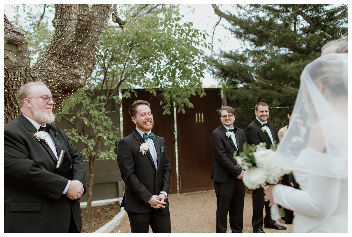 The groom stands at the altar with an emotional smile as he awaits his bride at an Artspace111 Fort Worth wedding. Twinkling string lights wrap around a large tree, creating a romantic outdoor setting. 