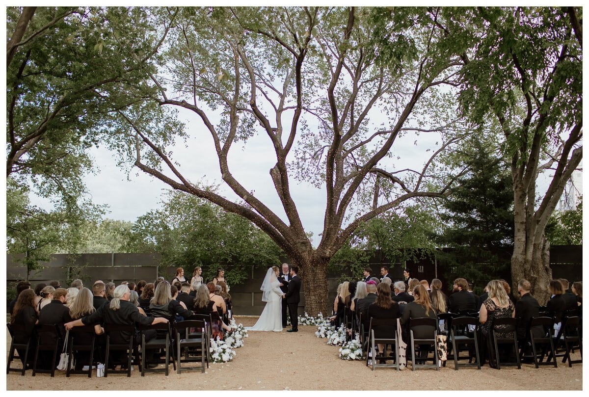 A romantic outdoor ceremony at an Artspace111 Fort Worth wedding, where the bride and groom stand beneath a grand tree wrapped in twinkling string lights. Surrounded by friends and family seated along a flower-lined aisle, they exchange vows in a beautifully intimate garden setting.