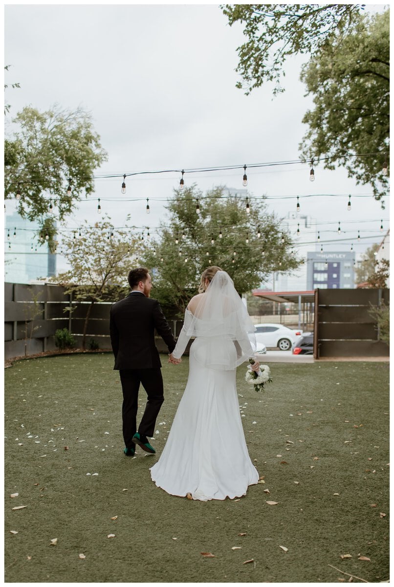 A newlywed couple walks hand in hand across the garden at their Artspace111 Fort Worth wedding. The bride, in a flowing white gown with an elegant veil, carries a bouquet of white flowers, while the groom, dressed in a classic black tuxedo, looks lovingly at her. Overhead, string lights add a romantic ambiance to the outdoor wedding venue, with the cityscape of Fort Worth visible in the background.