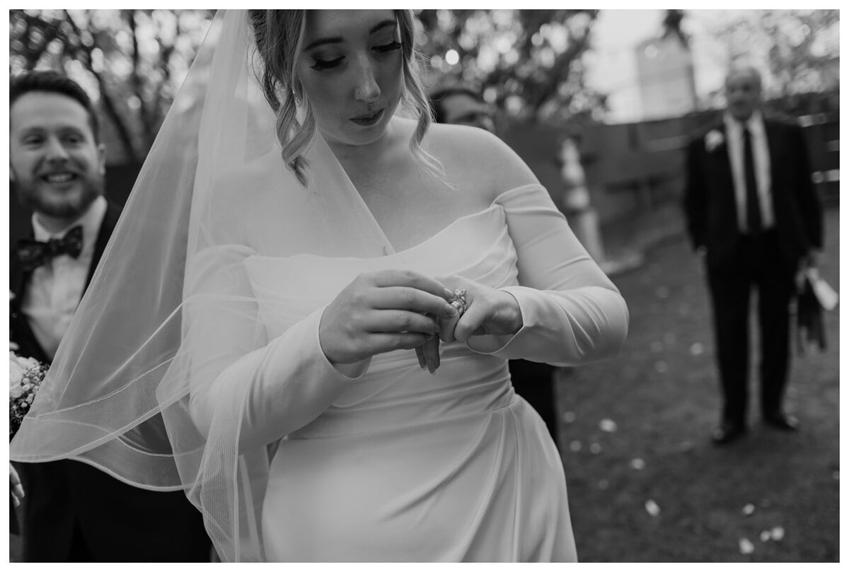 A bride delicately adjusts her wedding ring in this intimate black-and-white moment at her Artspace111 Fort Worth wedding. She wears an elegant off-the-shoulder gown with sheer and a flowing veil. In the blurred background, the groom smiles joyfully while wedding guests look on, capturing the emotion and beauty of this special day at the Fort Worth venue.