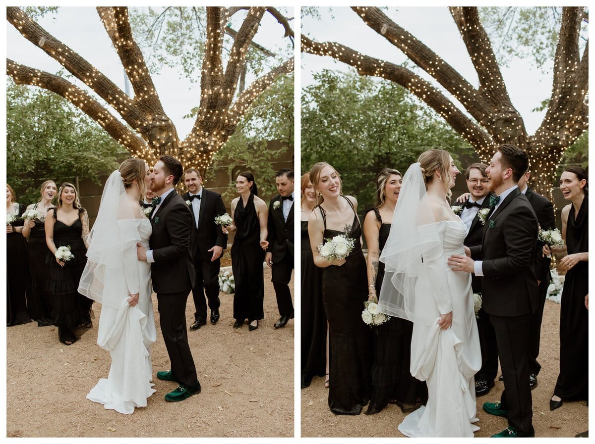 A joyful bride and groom share a kiss under twinkling tree lights during their Artspace111 Fort Worth wedding, surrounded by their cheering bridal party dressed in classic black. The couple's love and excitement shine in this candid moment at the beautiful outdoor venue, capturing the magic of their Fort Worth celebration.