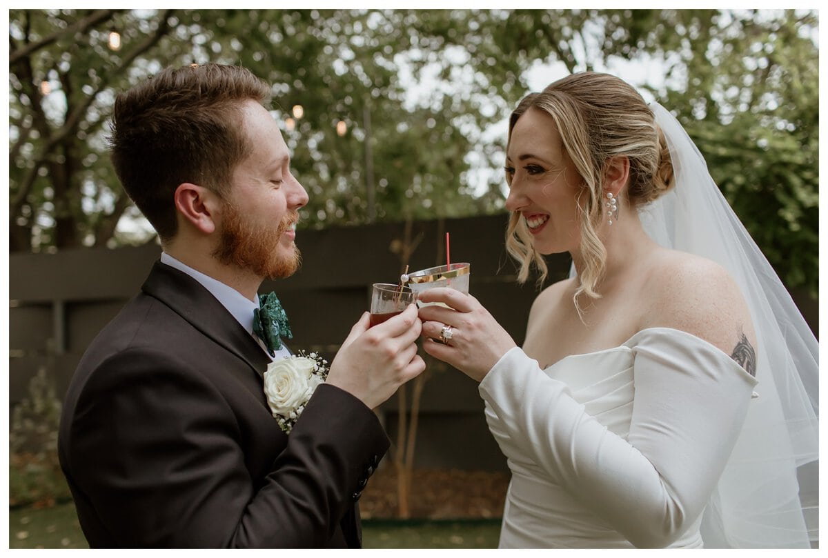 A joyful bride and groom share a celebratory toast at their Artspace111 Fort Worth wedding, smiling at each other with love. The bride stuns in an elegant off-the-shoulder white gown, while the groom looks dapper in a black tux with a green bow tie. The intimate garden setting with string lights adds to the romantic atmosphere of this special moment.