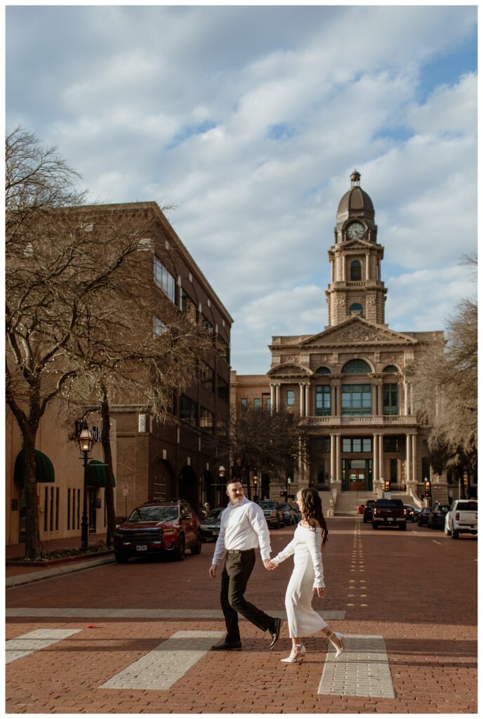 Engaged couple holding hands and crossing a brick street in downtown Fort Worth with the historic Tarrant County Courthouse in the background.