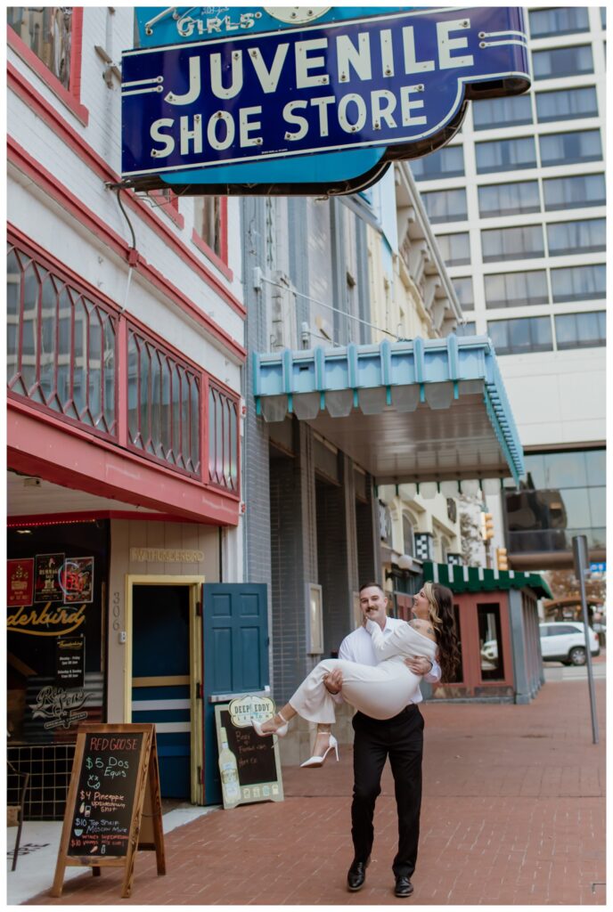 Engaged couple in downtown Fort Worth, with the groom-to-be playfully carrying his fiancée under the vintage 'Juvenile Shoe Store' sign, surrounded by historic architecture and city life.